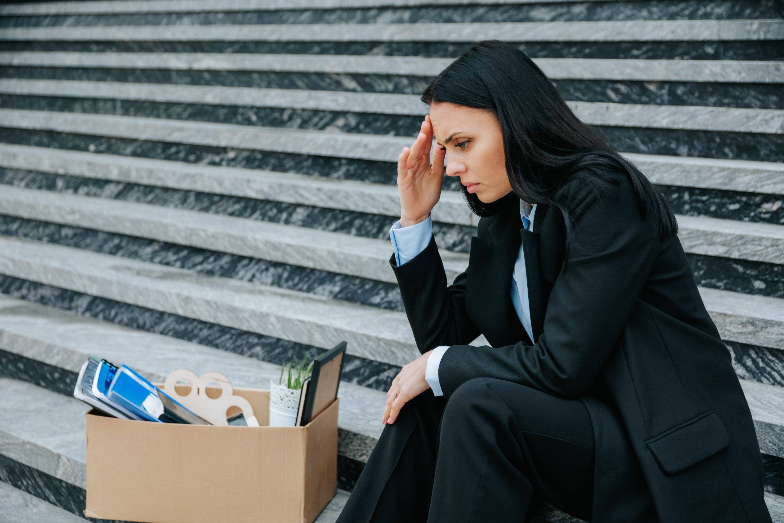 Female Looking Disappointed After Being Laid Off A powerful photo of a woman sitting on stairs, holding a cardboard box, representing the physical and emotional loss of her fired job and worklessness.
