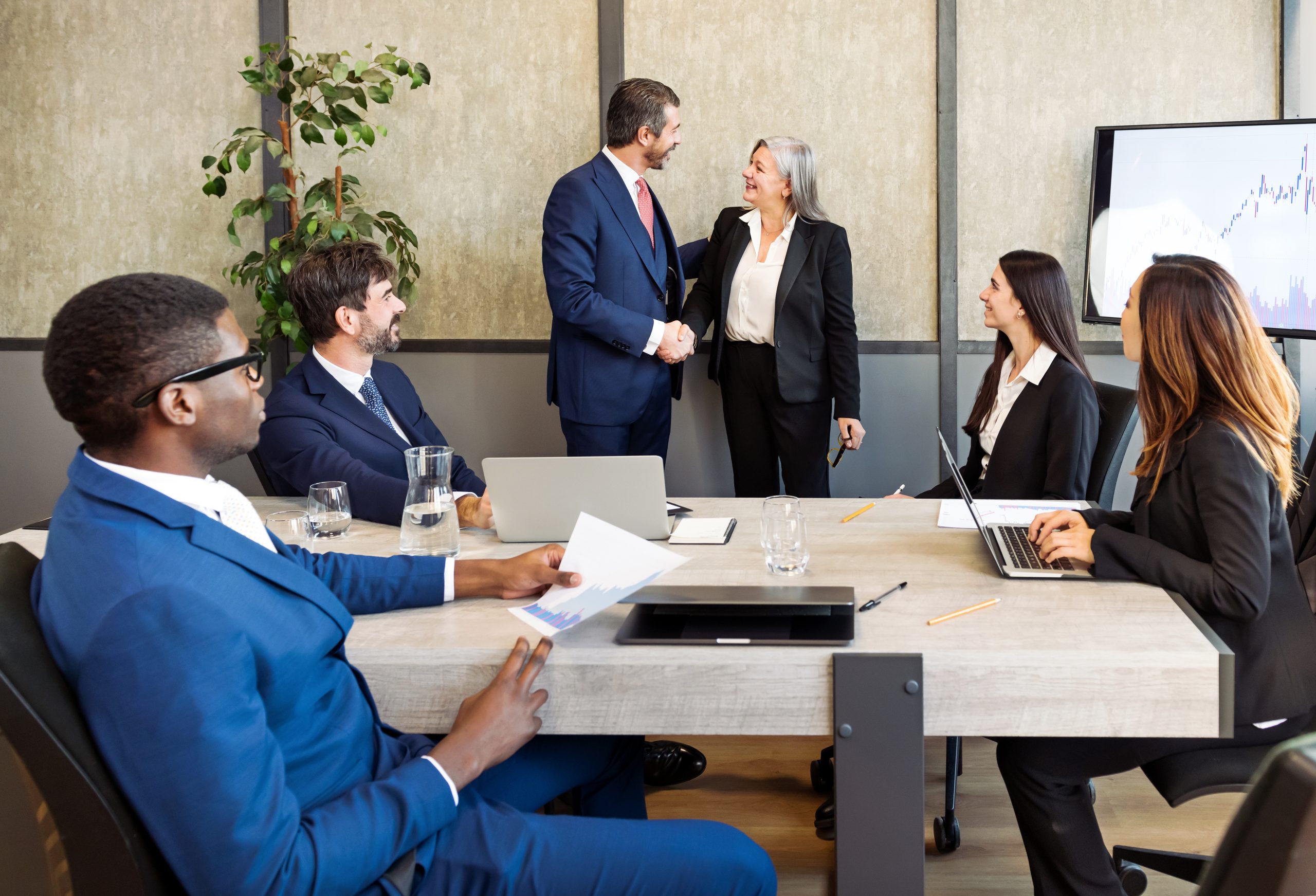 Group of diverse businesspeople in formal wear sitting at table and looking at coworkers shaking hands during business meeting in modern office