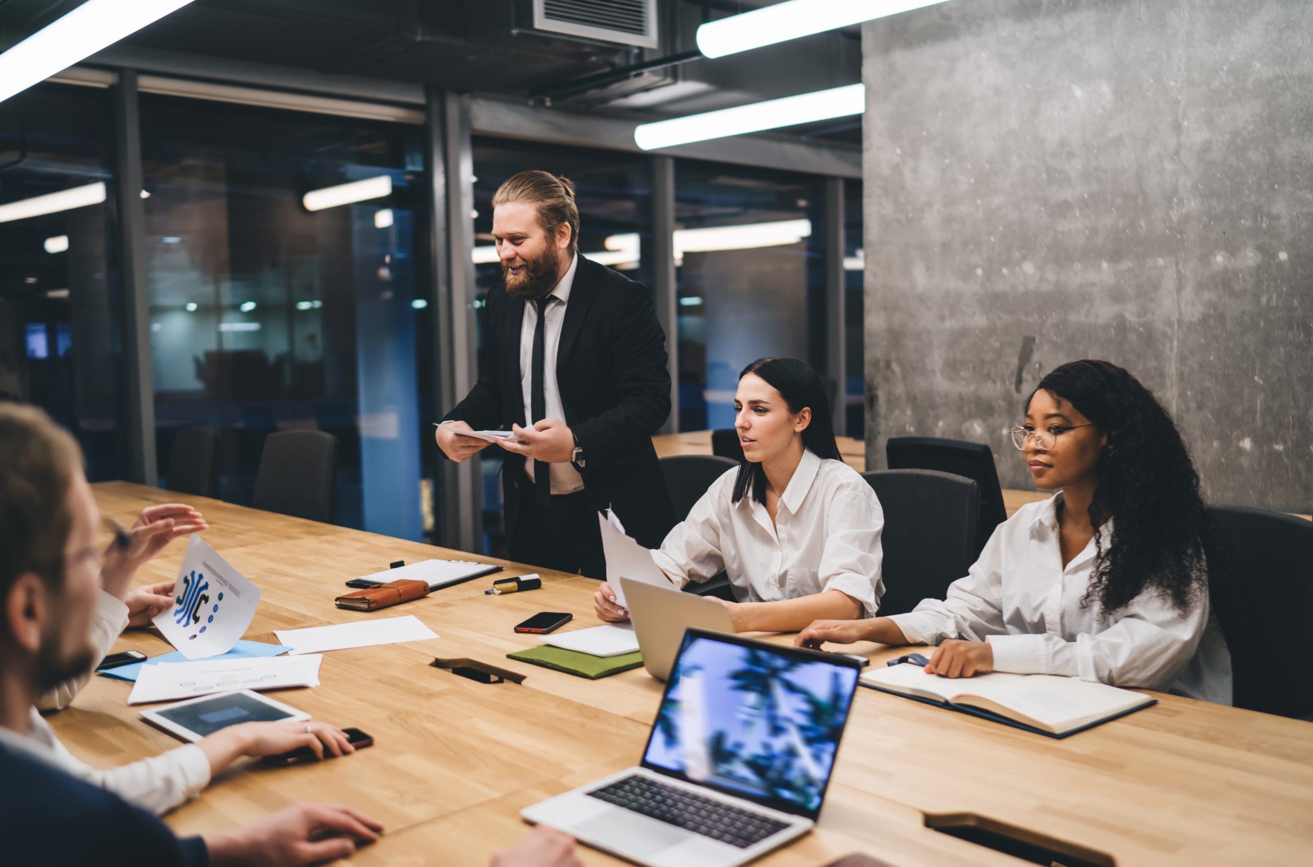 Multiethnic entrepreneurs in formal outfit sitting at table with documents and laptop while listening to colleague sharing business strategy in conference room