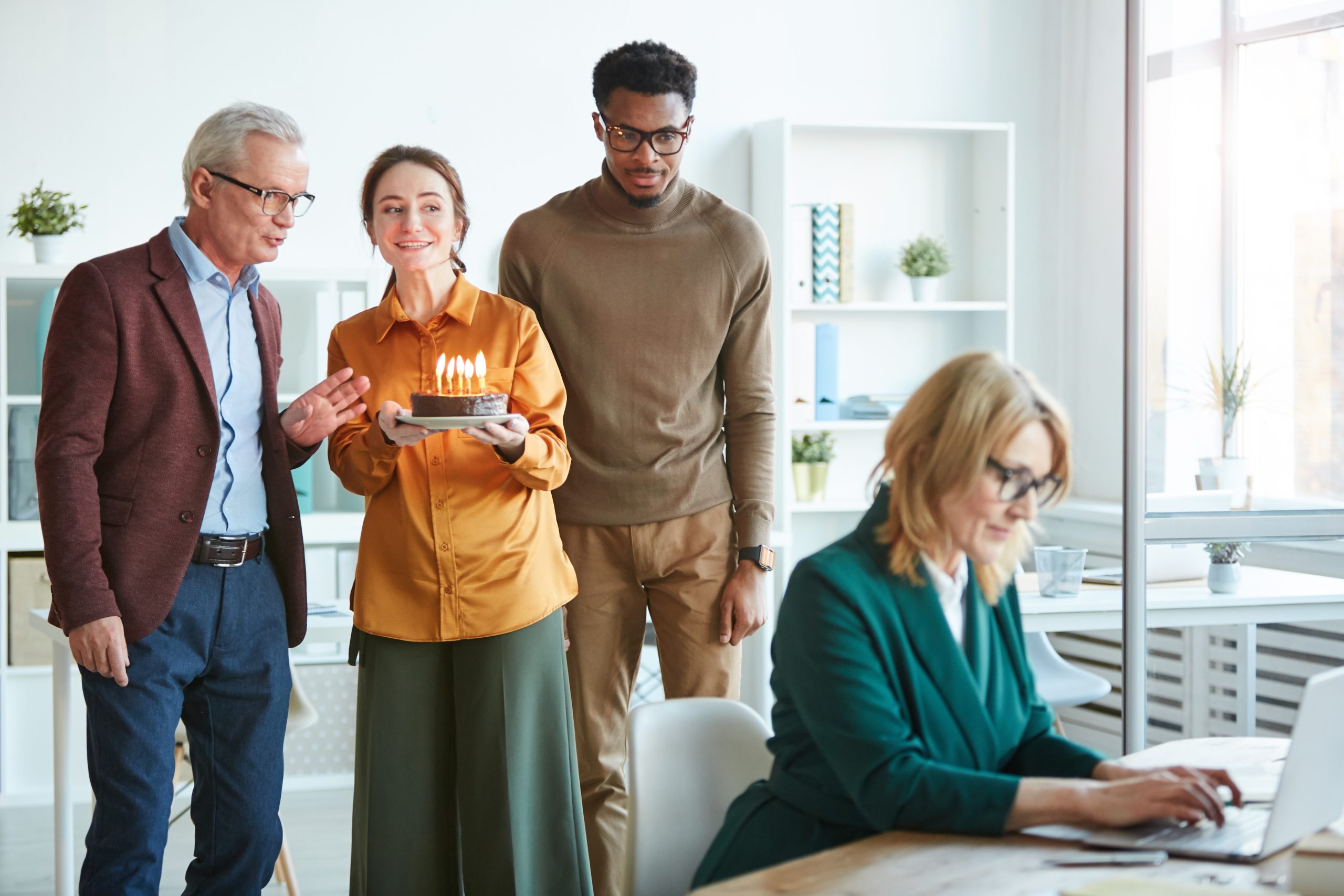 Business people holding cake with candles they are going to congratulate businesswoman who working at her table at office